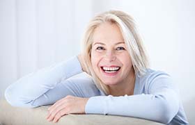 senior woman with dental implants in Aurora smiling and leaning over the back of a couch
