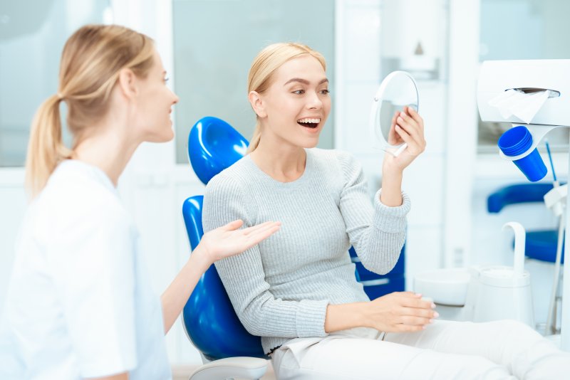 Woman admiring her smile in handheld mirror on blue dental chair while dentist talks to her